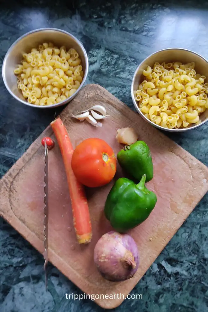 garlic, ginger, capsicum, carrots, tomatoes, and onions on the chopping board, and macaroni pasta in two bowls