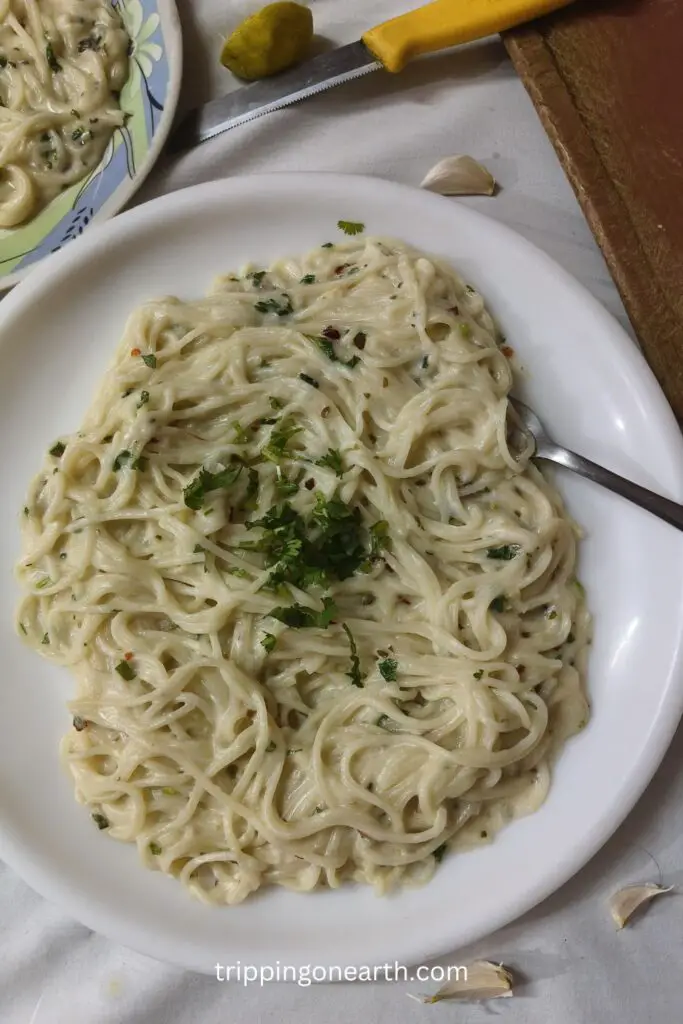 creamy lemon garlic pasta on a plate, garnished with coriander leaves, and lemon, garlic and knife cloves on the table beside it