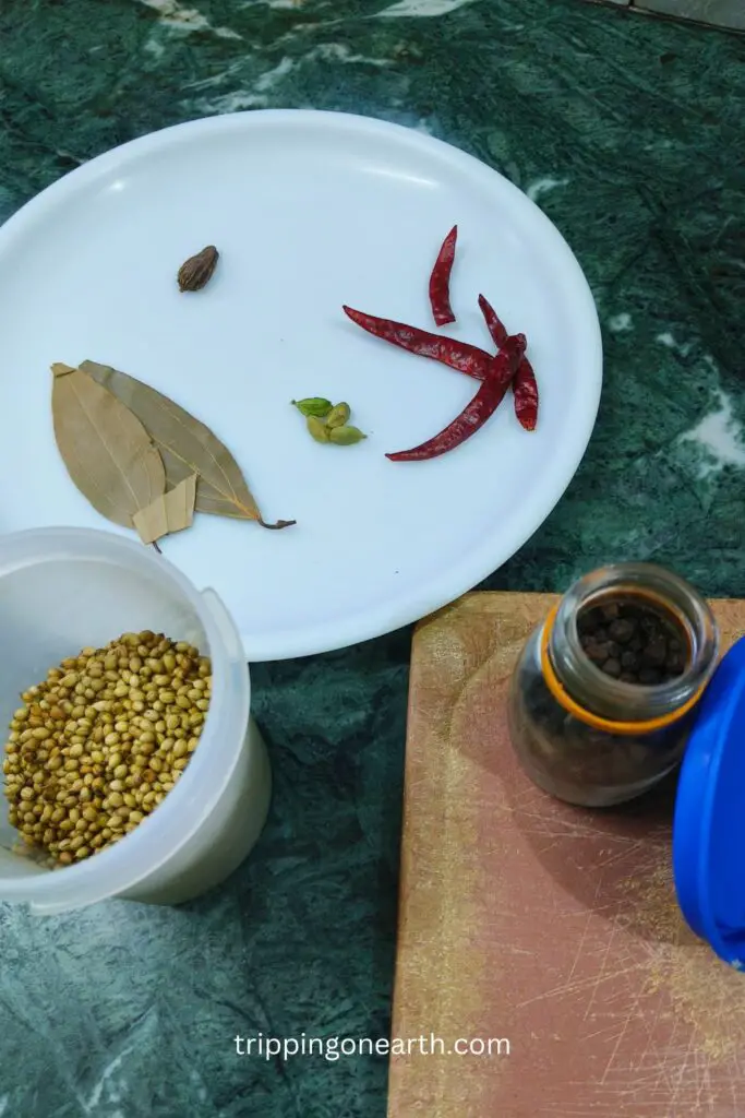 bay leaves, dried red chilies, black and green cardamoms on a white plate, and coriander seeds and black peppercorn in their respective containers