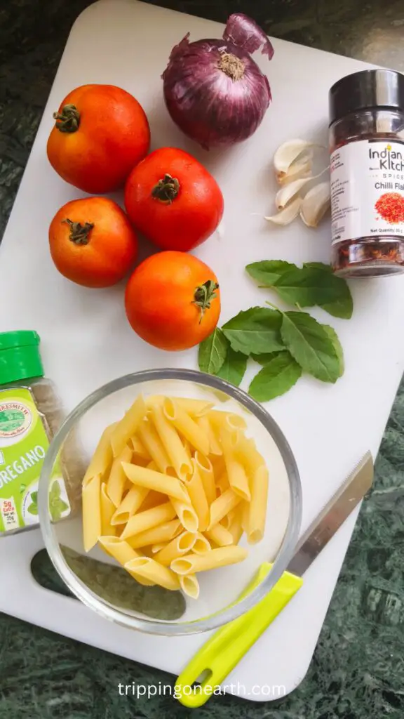 Ingredients on a white chopping board: Tomatoes, onion, garlic, penne pasta, red chili flakes, oregano, basil leaves
