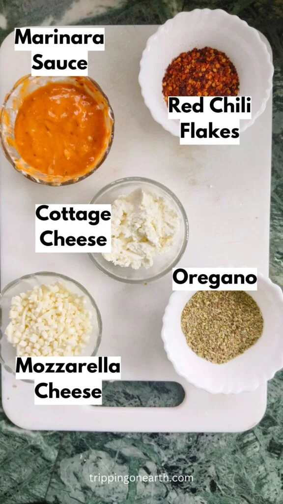 Ingredients laid out on a white chopping board: Marinara Sauce, red chili flakes, cottage cheese, shredded mozzarella cheese, and oregano