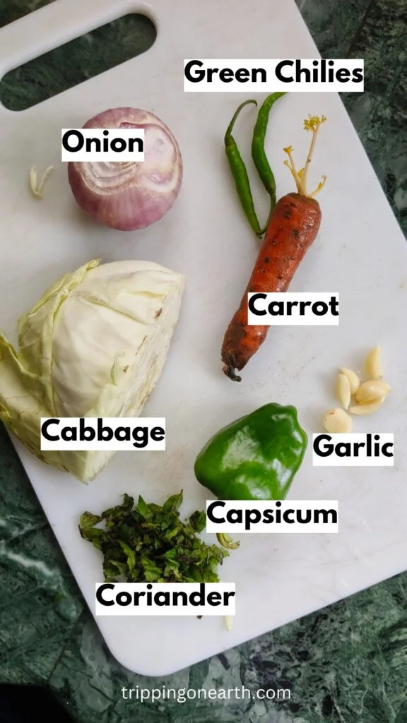 Ingredients laid out on a white chopping board: Green chilies, onion, capsicum, coriander, carrot, cabbage, and garlic