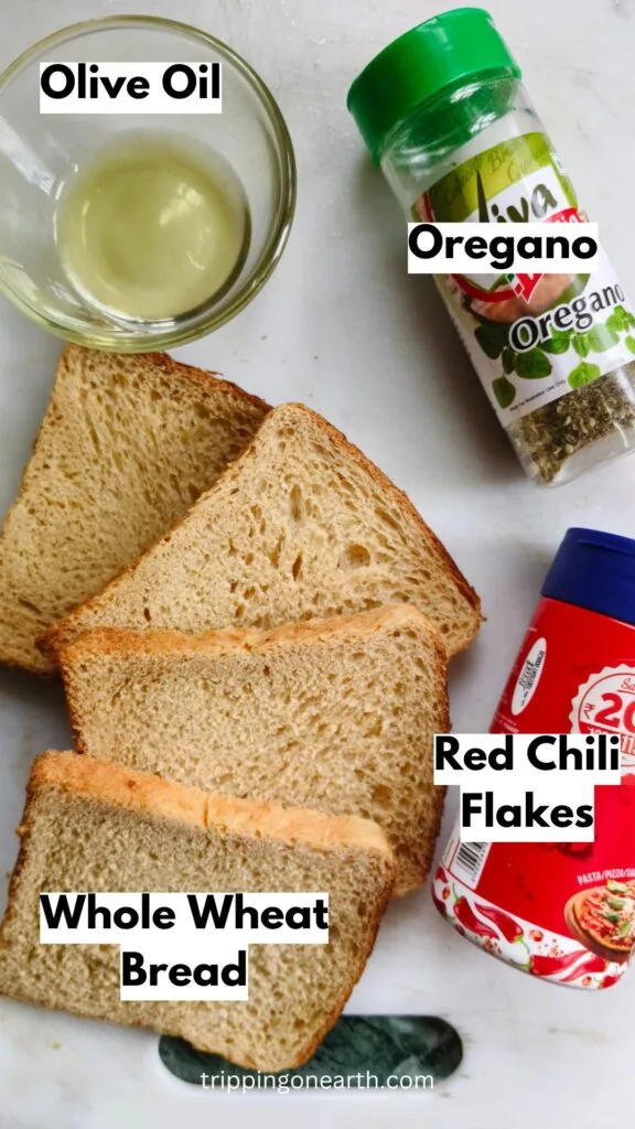 Ingredients laid out on a white cutting board- Olive oil, oregano, whole wheat bread, and red chili flakes