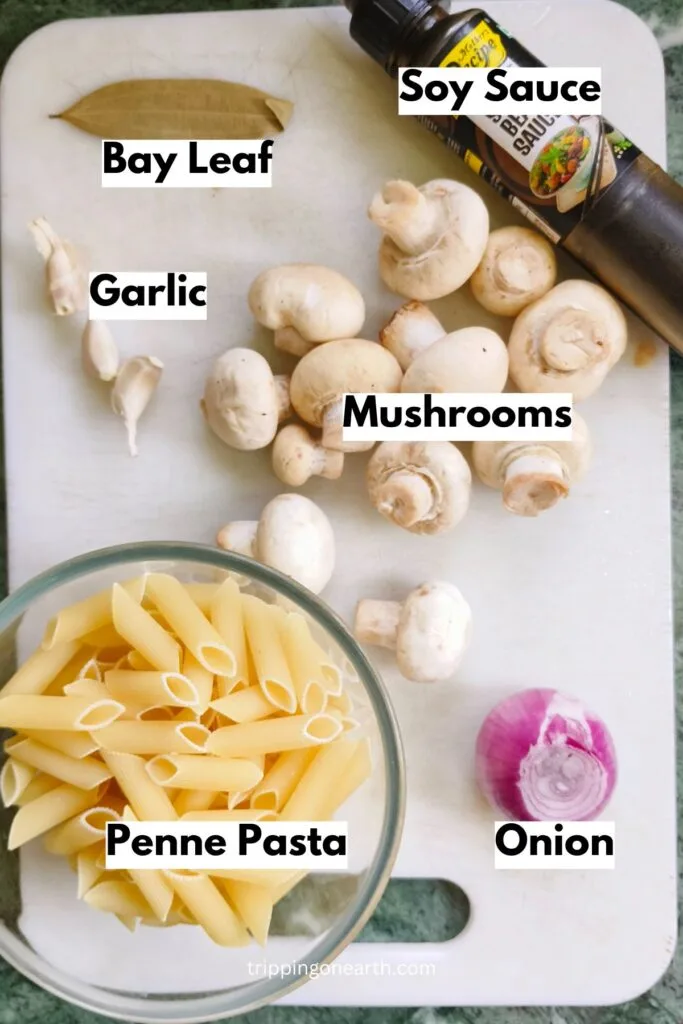 vegan mushroom stroganoff recipe ingredients- soy sauce, bay leaf, penne pasta, mushrooms, onion, and garlic laid on a white chopping board